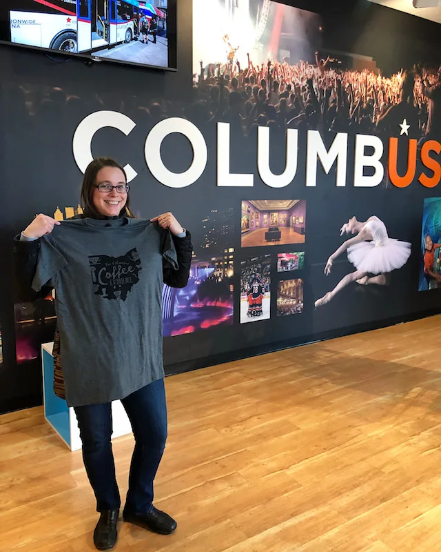 A woman holding up a Columbus Coffee Trail t-shirt at the Experience Columbus center in downtown Columbus, Ohio.