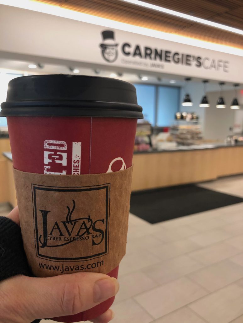 A cup of coffee held up in front of Carnegie's Cafe, a coffee shop inside the Main Library in downtown Columbus, Ohio.