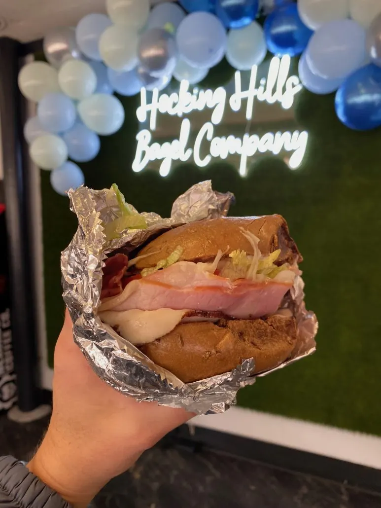 A bagel sandwich in front of a neon sign that says "Hocking Hills Bagel Company".