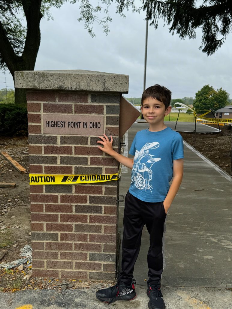 A boy standing next to a sign that says Highest Point in Ohio.